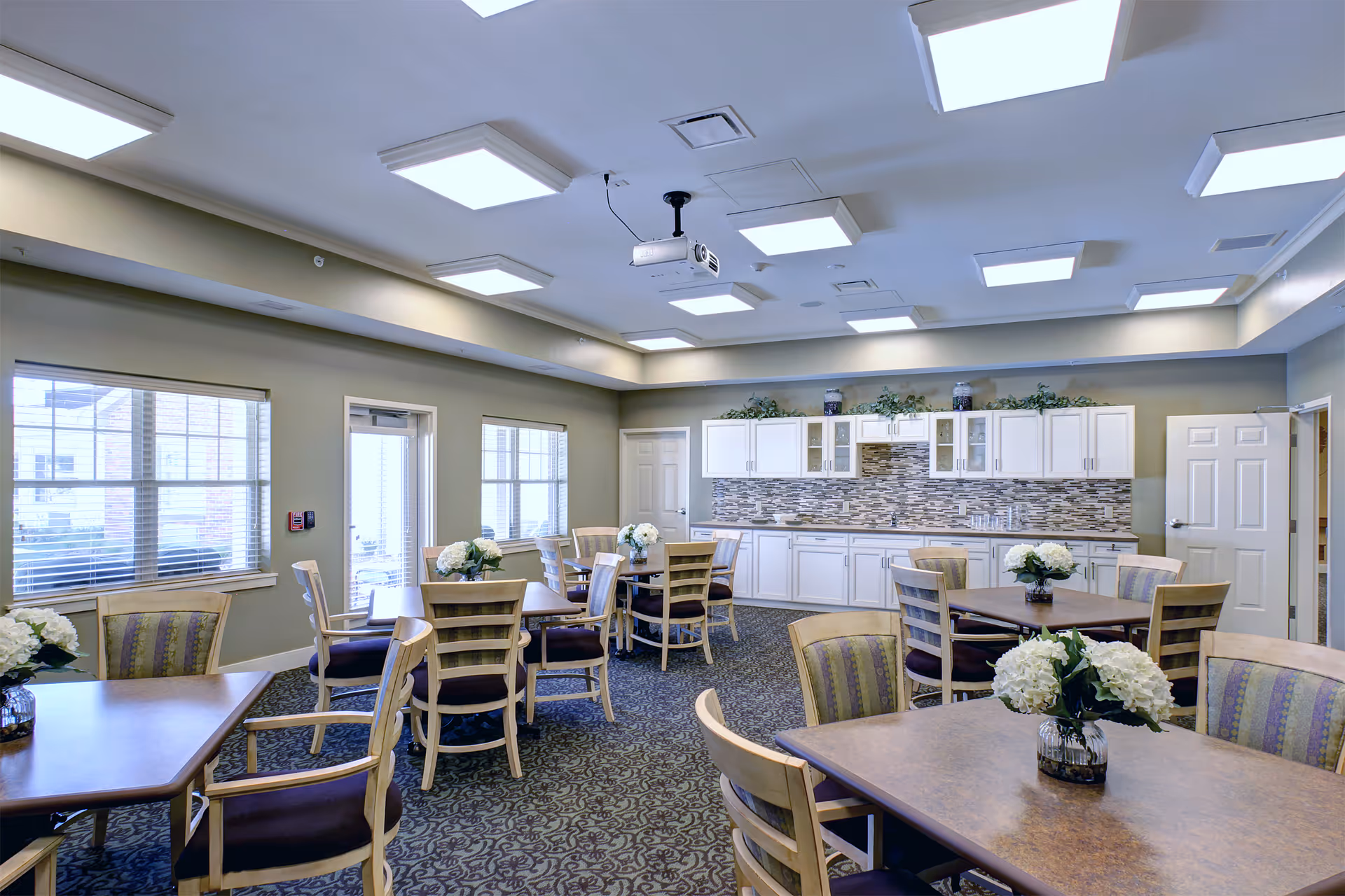 Well-lit communal dining room with multiple tables and chairs, floral centerpieces, and a kitchenette with white cabinets and a tiled backsplash along the back wall.
