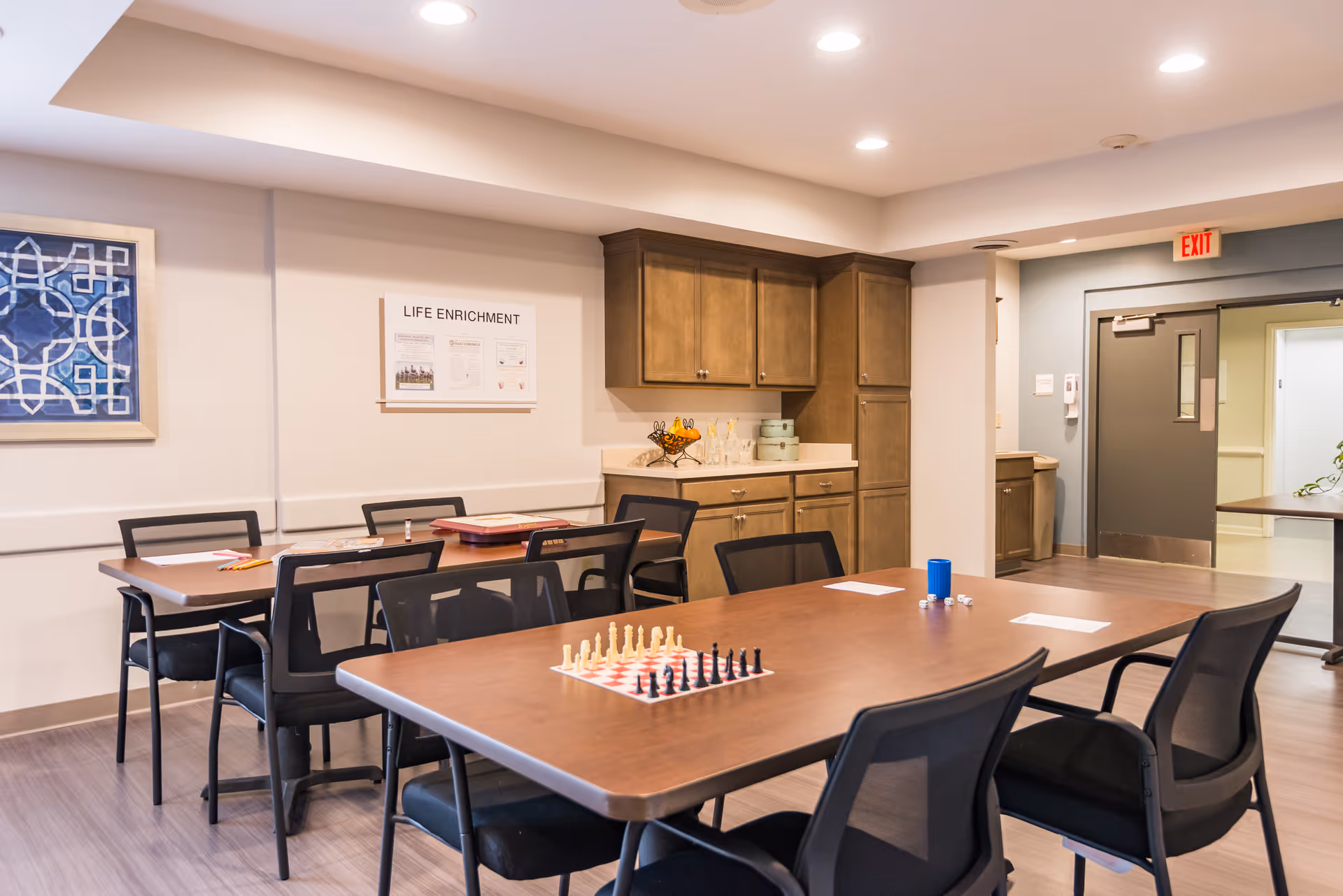 A well-lit room with two wooden tables surrounded by black chairs. One table has a chessboard with chess pieces set up, papers, dice, and a blue cup. The other table has some papers and writing materials. There are wooden cabinets and a countertop with a fruit basket and glassware against the wall. A framed artwork and a 'Life Enrichment' bulletin board are mounted on the wall. The room has a light-colored floor and ceiling with recessed lighting, and a gray door with an exit sign above it is visible in the background.