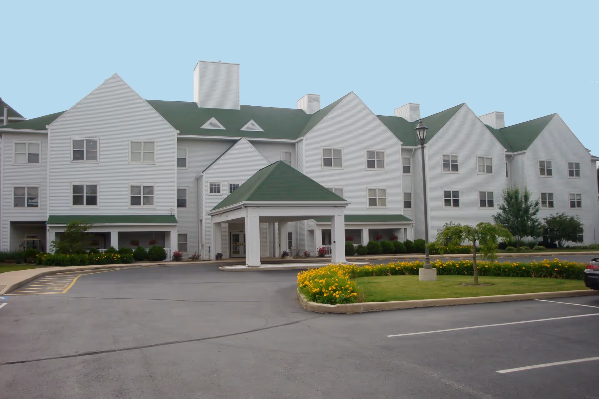Exterior view of a large, white, multi-story assisted living community building with green roofs and a covered entrance. There is a parking lot in front with some yellow flowers and small trees in landscaped areas.