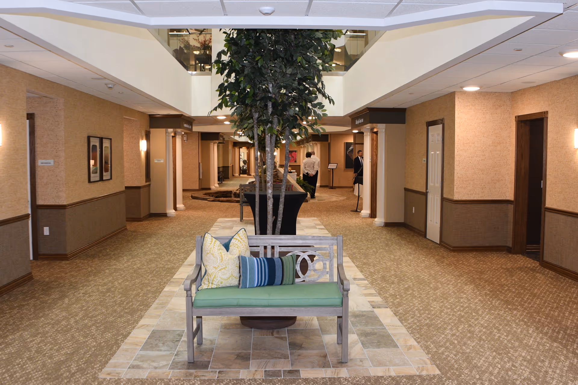 Spacious indoor lobby corridor with a bench, cushions, and a central potted tree leading to hallways.