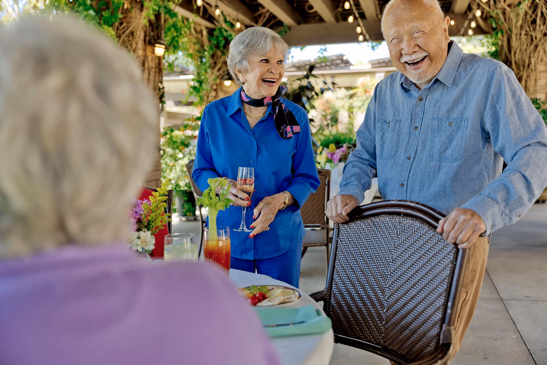 Three elderly people socializing outdoors under a covered patio. One woman in a blue shirt is holding a glass of drink and smiling, a man in a blue shirt is standing and holding a chair while laughing, and another person with gray hair is seated with their back to the camera. The setting includes plants and flowers, creating a warm and inviting atmosphere.