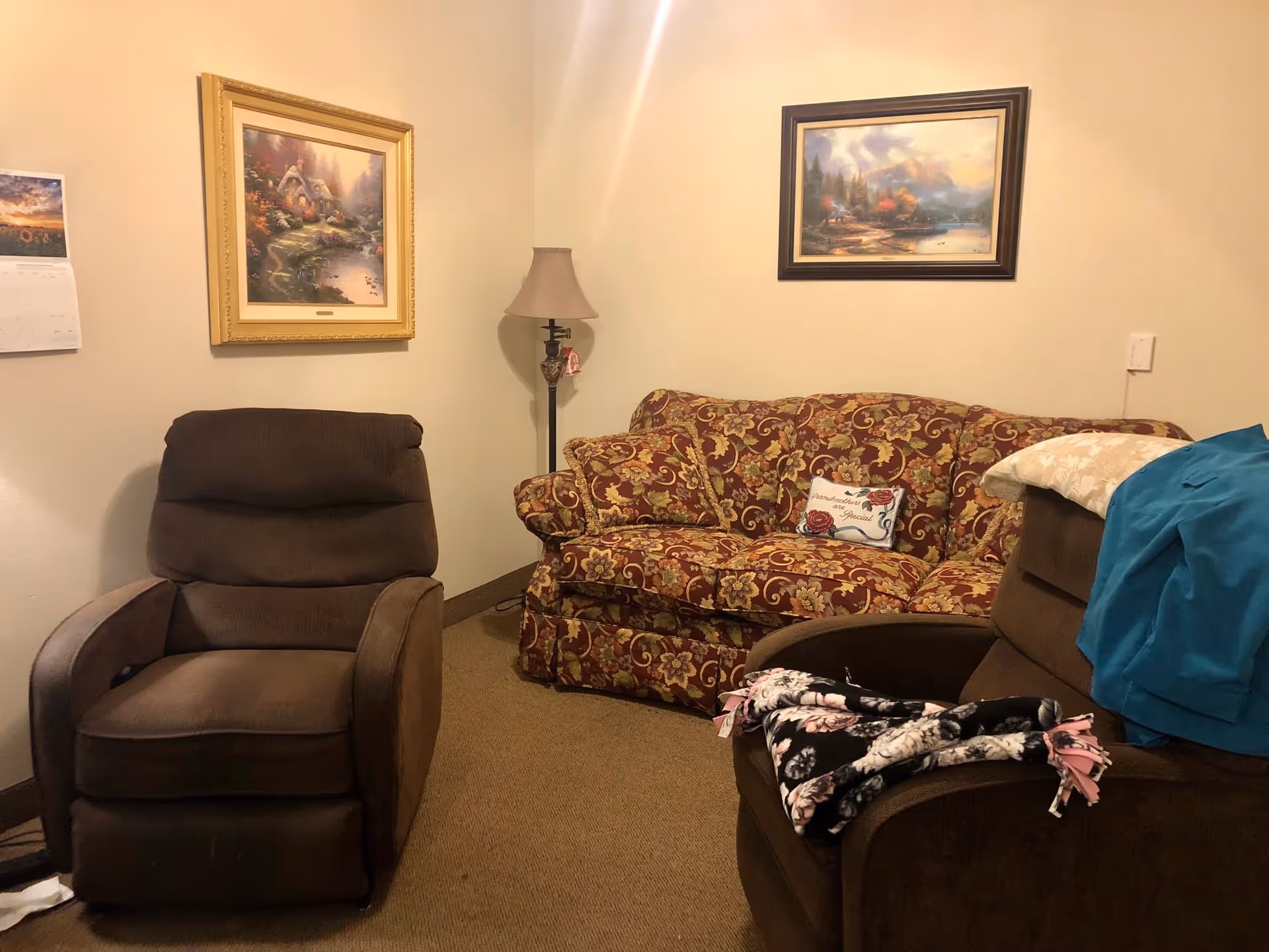 A cozy living room with two brown recliner chairs and a floral patterned sofa. The sofa has a decorative pillow that reads 'Grandmother's are Special.' There are two framed landscape paintings on the beige walls and a floor lamp in the corner. A calendar is partially visible on the left wall.