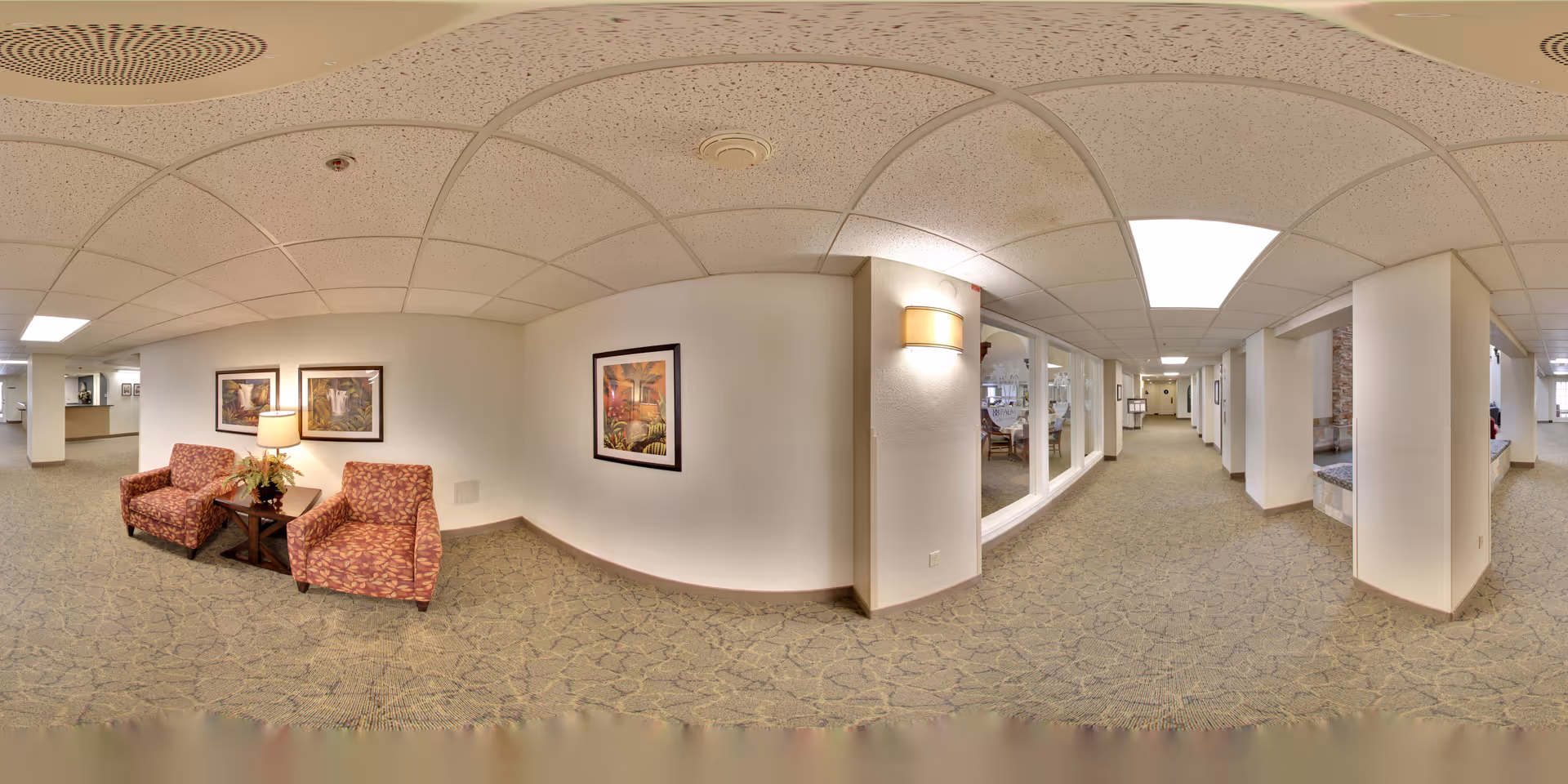 Well-lit carpeted hallway in a senior living facility with two patterned chairs and a side table under framed artwork beside a long corridor and windows into common areas.