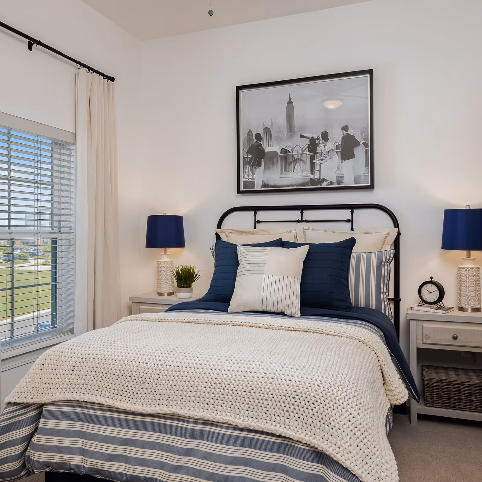 A neatly made bedroom with a black metal bedframe, navy and cream bedding, two bedside tables with blue lamps, and a framed black-and-white photo above the bed.