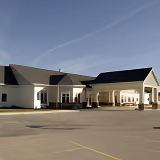 Exterior view of a single-story building with a covered entrance and a large paved parking area under a clear blue sky.