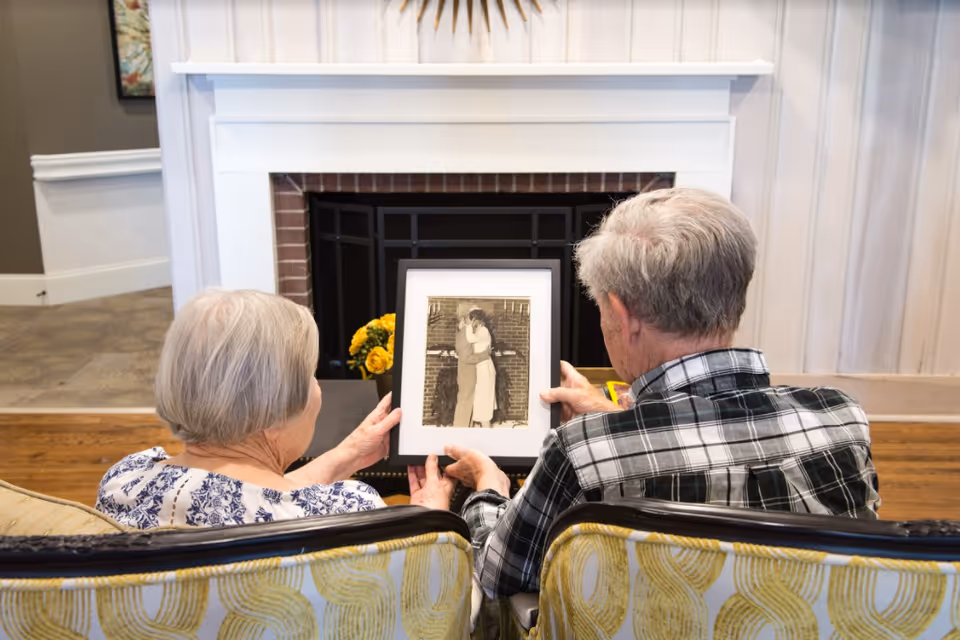 An elderly couple sitting on a patterned couch in a living room, holding and looking at a framed black-and-white photograph of a couple embracing. Behind them is a white fireplace with a black screen and a vase of yellow flowers on the hearth.