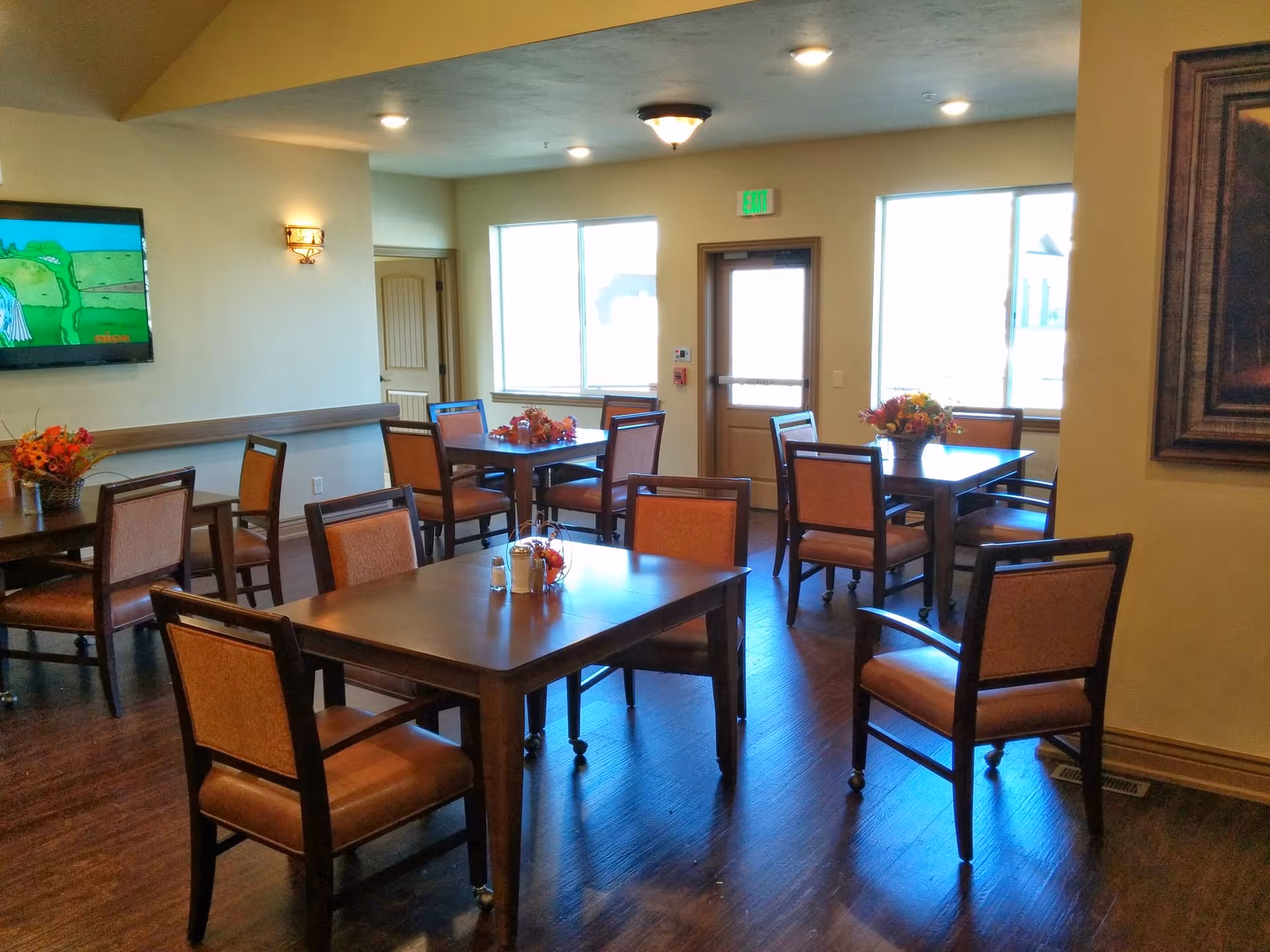 A dining room with several wooden tables and chairs arranged neatly. Each table has a small floral centerpiece. The room has wooden flooring, two large windows letting in natural light, a door with an exit sign above it, and a wall-mounted TV displaying a cartoon. There is a framed painting on the right wall.