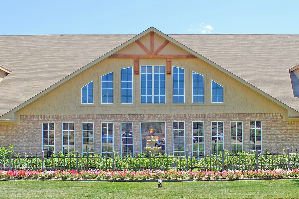 Front exterior of a brick-and-siding building with a large gabled roof, rows of tall windows, a fenced flowerbed and manicured lawn.
