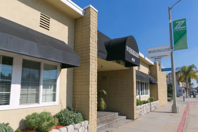 Exterior view of Shoreline Healthcare Center building with beige brick walls, black awnings over windows and entrance, and a sidewalk with street signs and palm trees in the background.