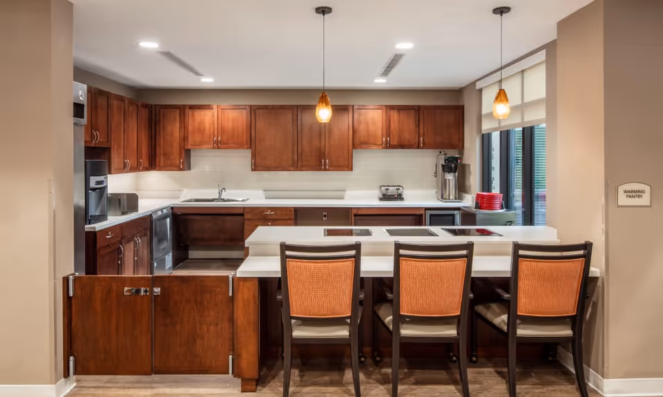 Modern communal kitchen with a white island, four chairs, wooden cabinets and pendant lights.