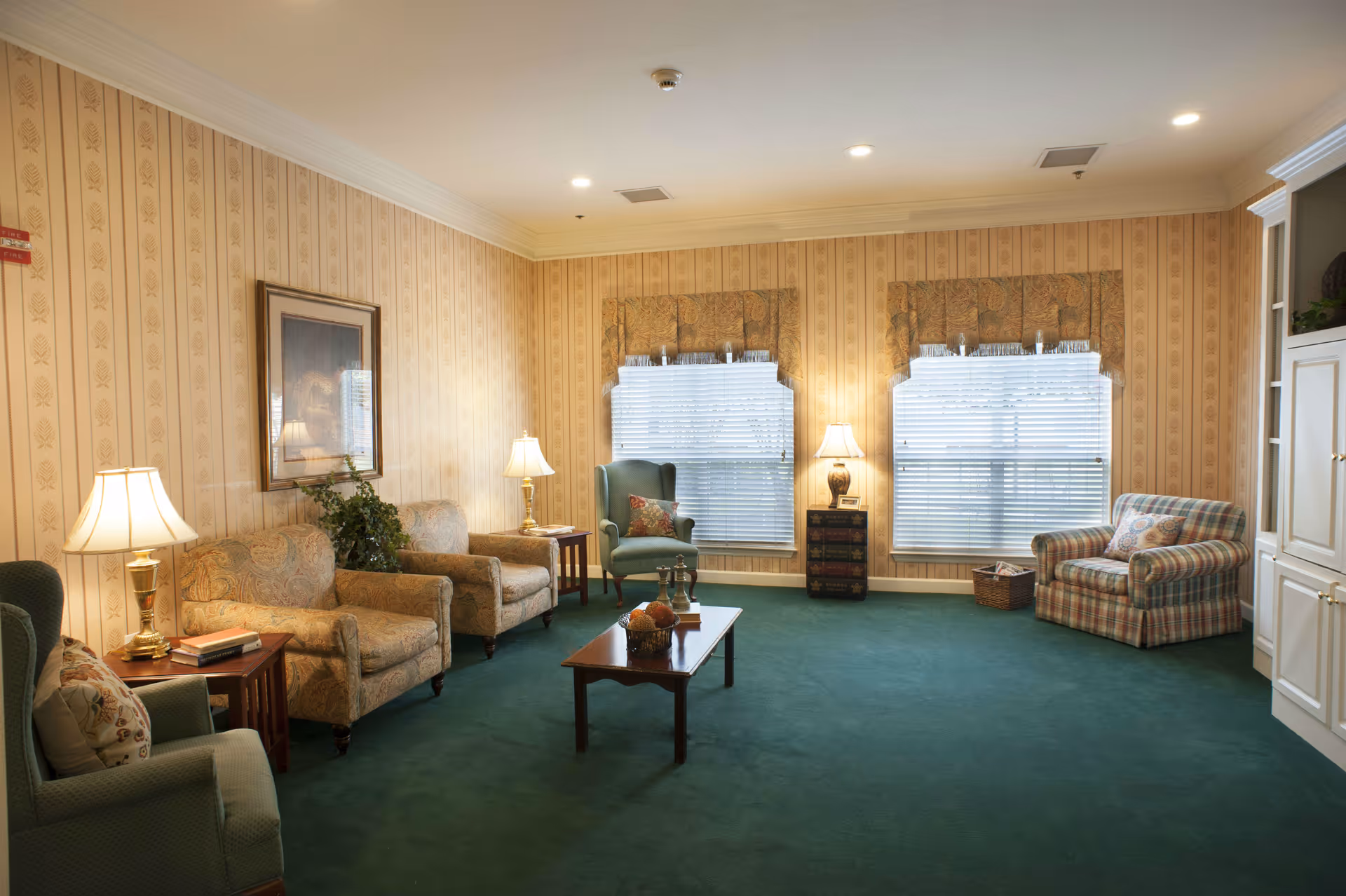 A cozy living room with green carpet and beige patterned wallpaper. The room features multiple upholstered chairs and sofas in various patterns, a wooden coffee table with decorative items, two large windows with blinds and valances, several table lamps providing warm lighting, a framed picture on the wall, and a white built-in cabinet on the right side.
