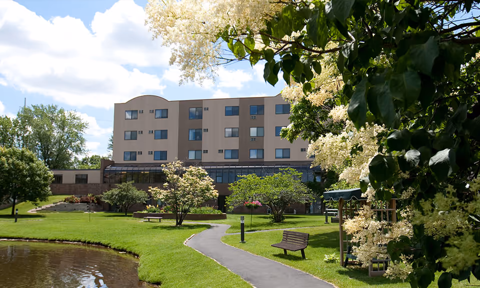 Front view of a multi-story senior living building with a landscaped lawn, walking path, pond, benches, and flowering trees.