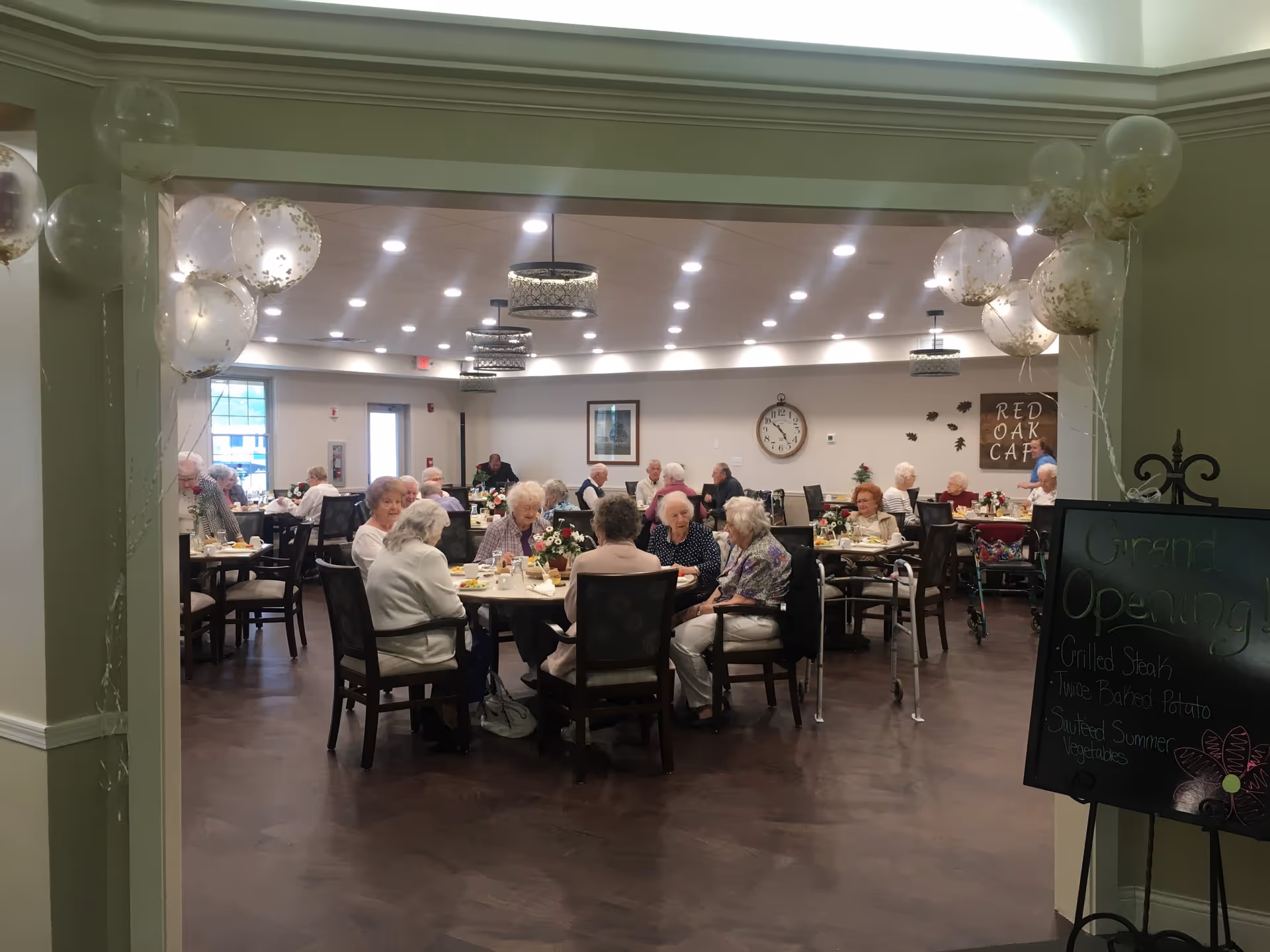 A dining room in a senior living facility with elderly residents seated at tables enjoying a meal. The room is decorated with balloons and a chalkboard sign announcing a grand opening with menu items. The walls have a clock and a sign that reads 'Red Oak Cafe'.