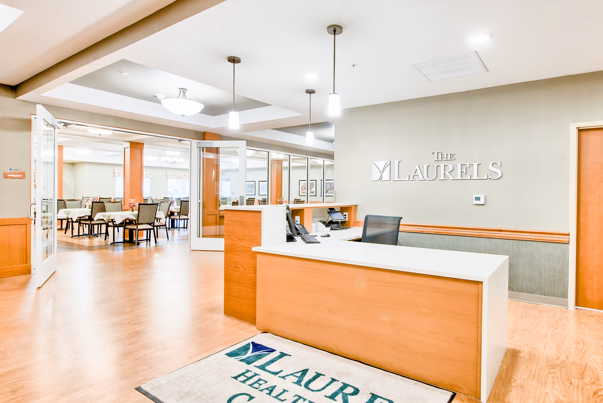 Reception desk area with a chair, computer, and phone in front of a wall with The Laurels sign. To the left, double glass doors open into a dining room with tables and chairs arranged neatly. The floor is wooden, and the space is well-lit with ceiling lights and pendant lamps.