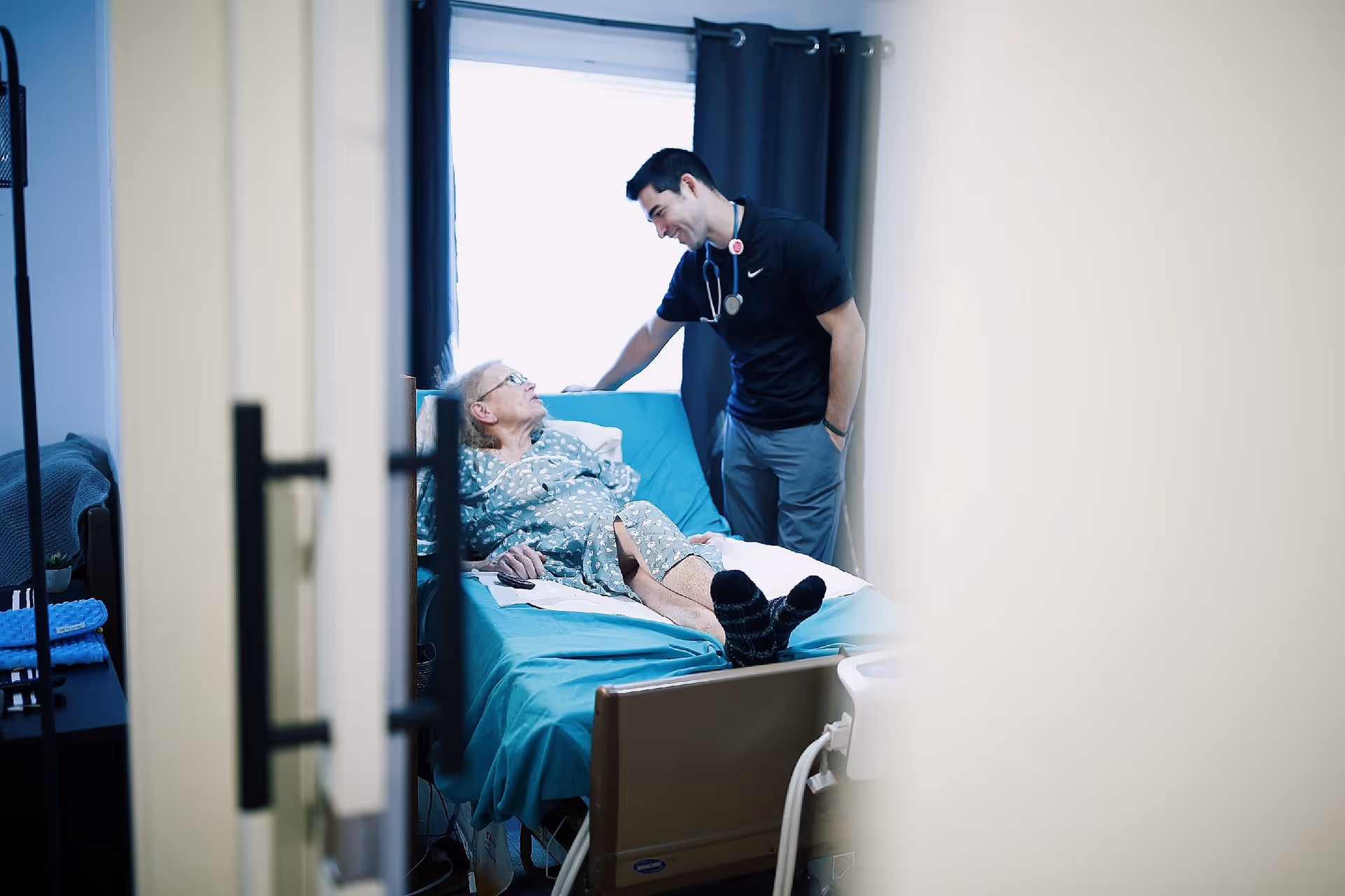 An elderly woman lying in a hospital bed wearing a hospital gown, looking up and smiling at a male healthcare professional standing beside her. The healthcare professional is wearing a black shirt with a stethoscope around his neck and is leaning slightly towards the woman in a caring manner. The scene is viewed through a partially open door with a window in the background covered by dark curtains.