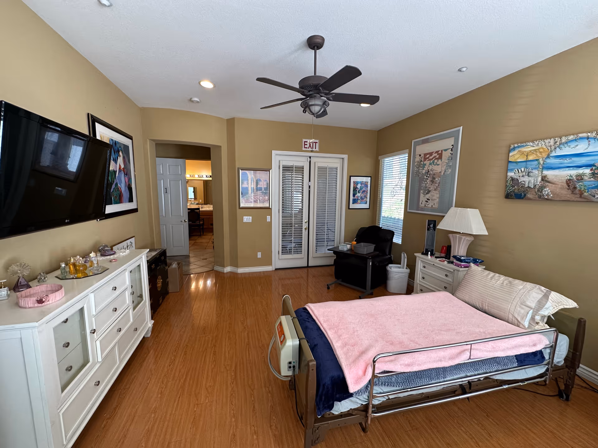 Spacious resident bedroom with a hospital-style bed covered by a pink blanket, dresser with TV, armchair, ceiling fan, and double doors.