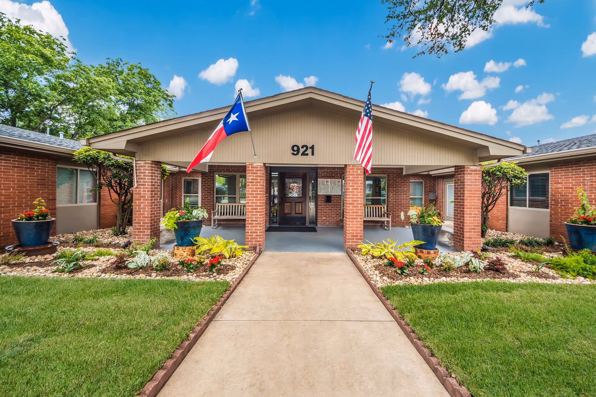 Front entrance of a single-story brick building with the number 921 above the door. Two flags, the Texas state flag and the United States flag, are displayed on poles attached to the building. There are landscaped garden beds with plants and flowers on either side of the concrete walkway leading to the entrance. The sky is blue with scattered clouds.
