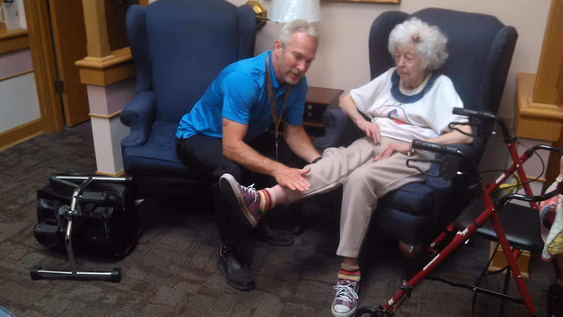 A staff member kneels to examine an elderly woman’s leg as she sits in a lounge chair with a walker nearby.