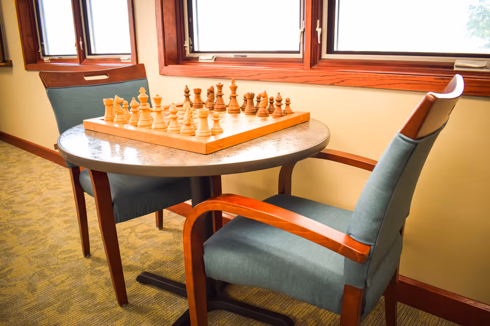 A small round table with a wooden chessboard set up for a game, placed near windows with wooden frames. Two cushioned chairs with wooden armrests are positioned on either side of the table in a well-lit room.
