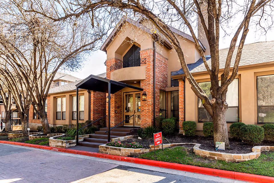 Front entrance of a retirement community building with a brick facade, covered steps, trees and landscaped beds.
