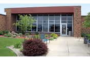 Front entrance of a brick building with a paved walkway, benches, shrubs, and large glass doors.
