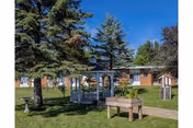 Grassy courtyard featuring a white gazebo, picnic table and raised planter surrounded by pine trees in front of a single-story brick building under a blue sky.