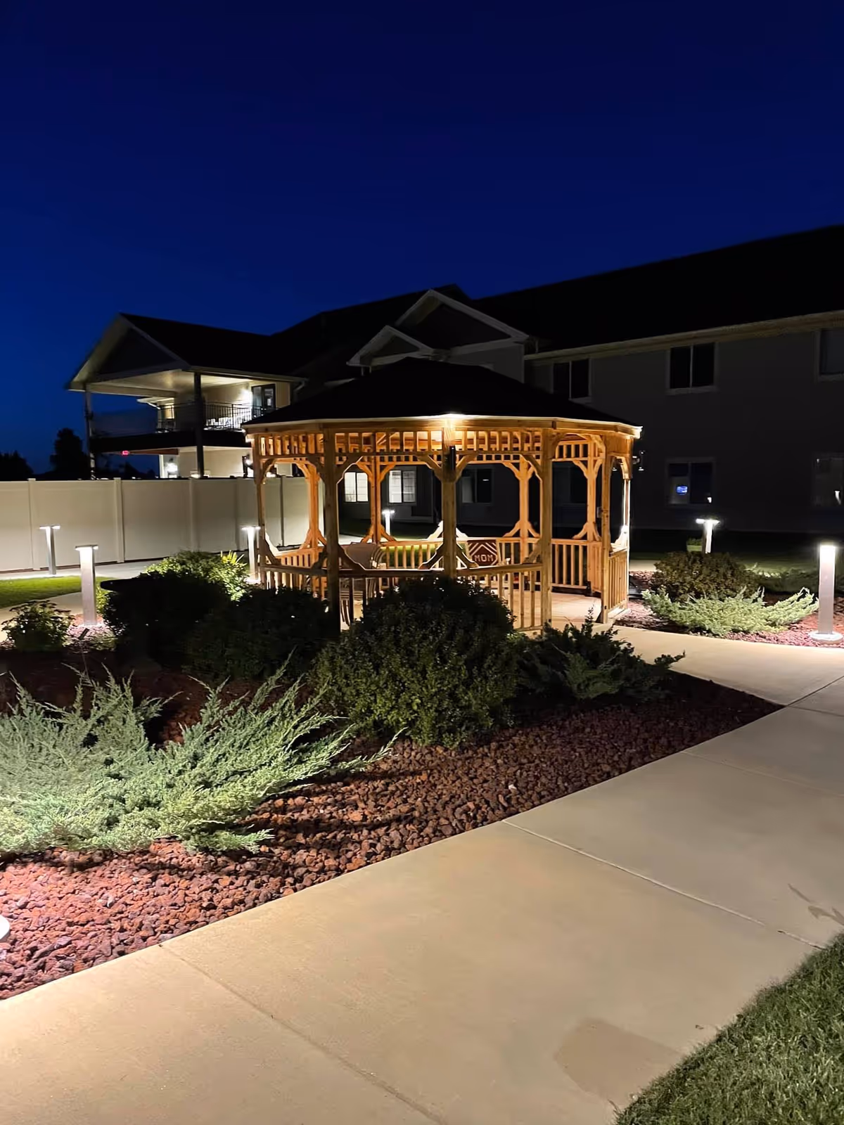 A wooden gazebo illuminated by lights at night, surrounded by landscaped bushes and a paved walkway. In the background, there is a two-story building with balconies and windows, and a white fence enclosing the area.