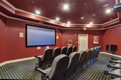 A small theater room with two rows of black cushioned seats facing a large blank projection screen mounted on a red wall. The room has recessed ceiling lights and patterned carpet flooring.