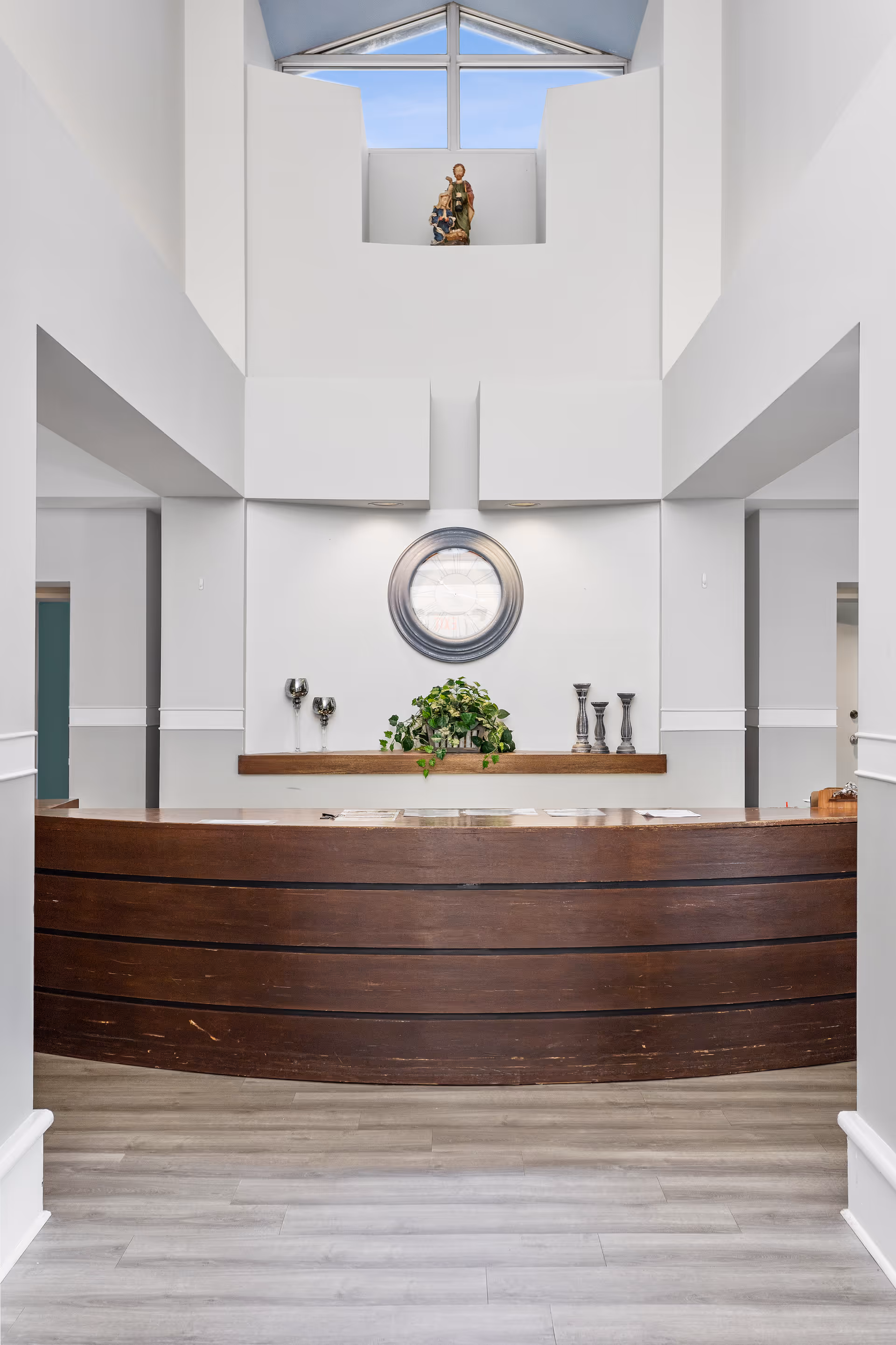 Reception area with a curved wooden desk, a round mirror on the wall, decorative candle holders, and a green plant. Above the wall is a small statue displayed near a triangular window showing a blue sky.