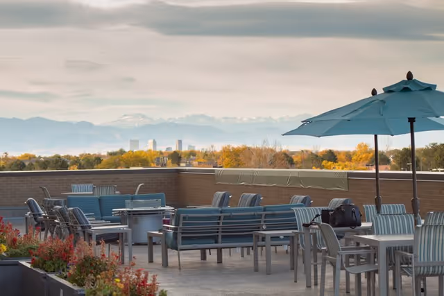 Outdoor rooftop patio area with multiple seating arrangements including chairs and benches, a large umbrella, and a scenic view of distant mountains and a city skyline under a cloudy sky.