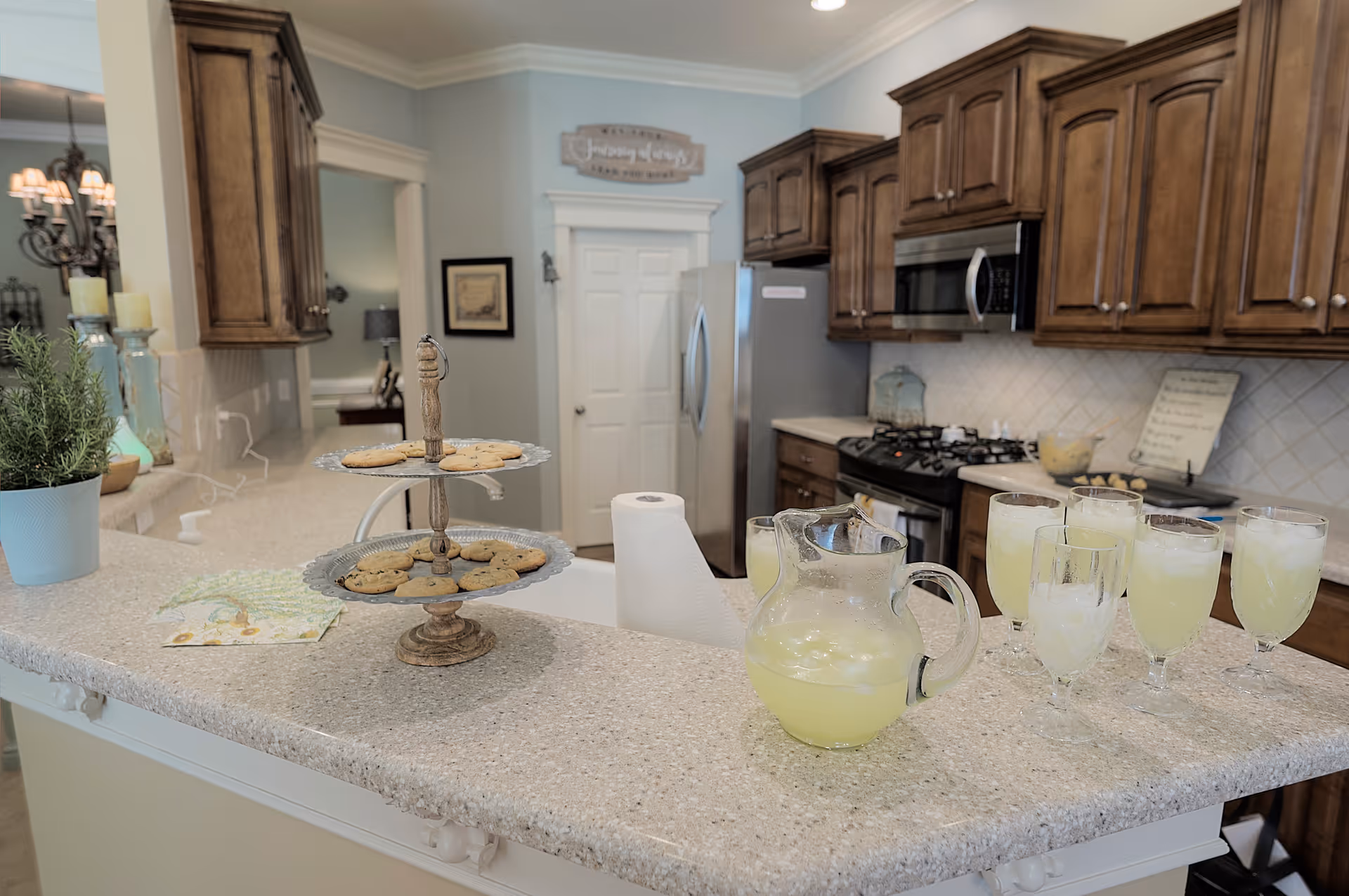 A kitchen with wooden cabinets, a stainless steel refrigerator, microwave, and gas stove. On the countertop, there is a two-tiered tray with cookies, a pitcher and glasses filled with lemonade, a roll of paper towels, and a potted plant. The kitchen has light-colored walls and a tiled backsplash.