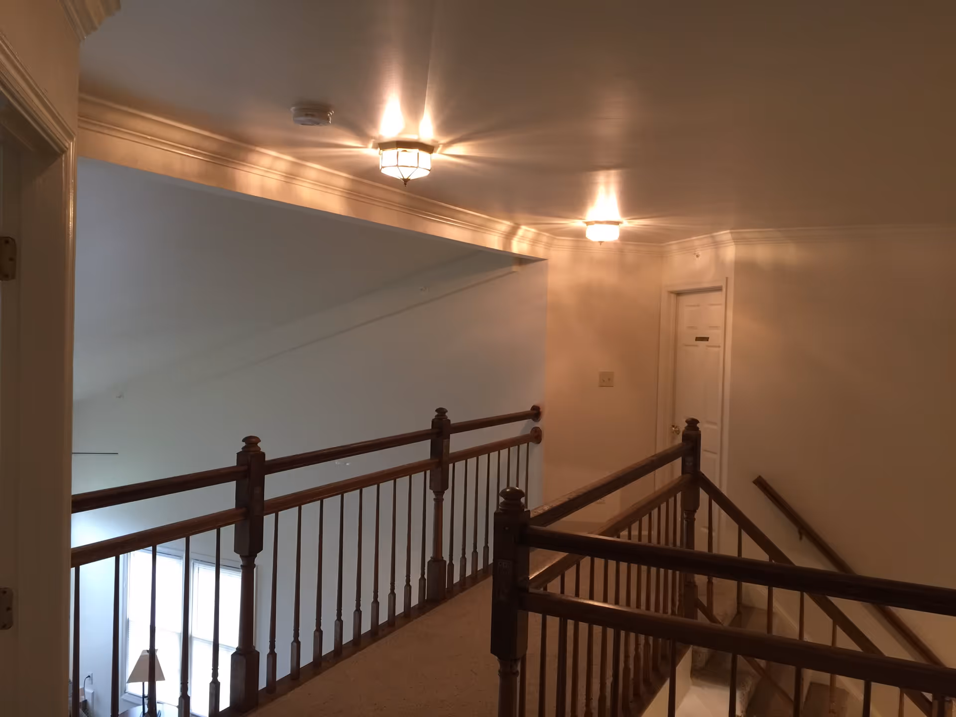 Interior view of a hallway with wooden railings and beige walls, illuminated by ceiling lights. A door is visible at the end of the hallway, and a staircase descends to a lower level.