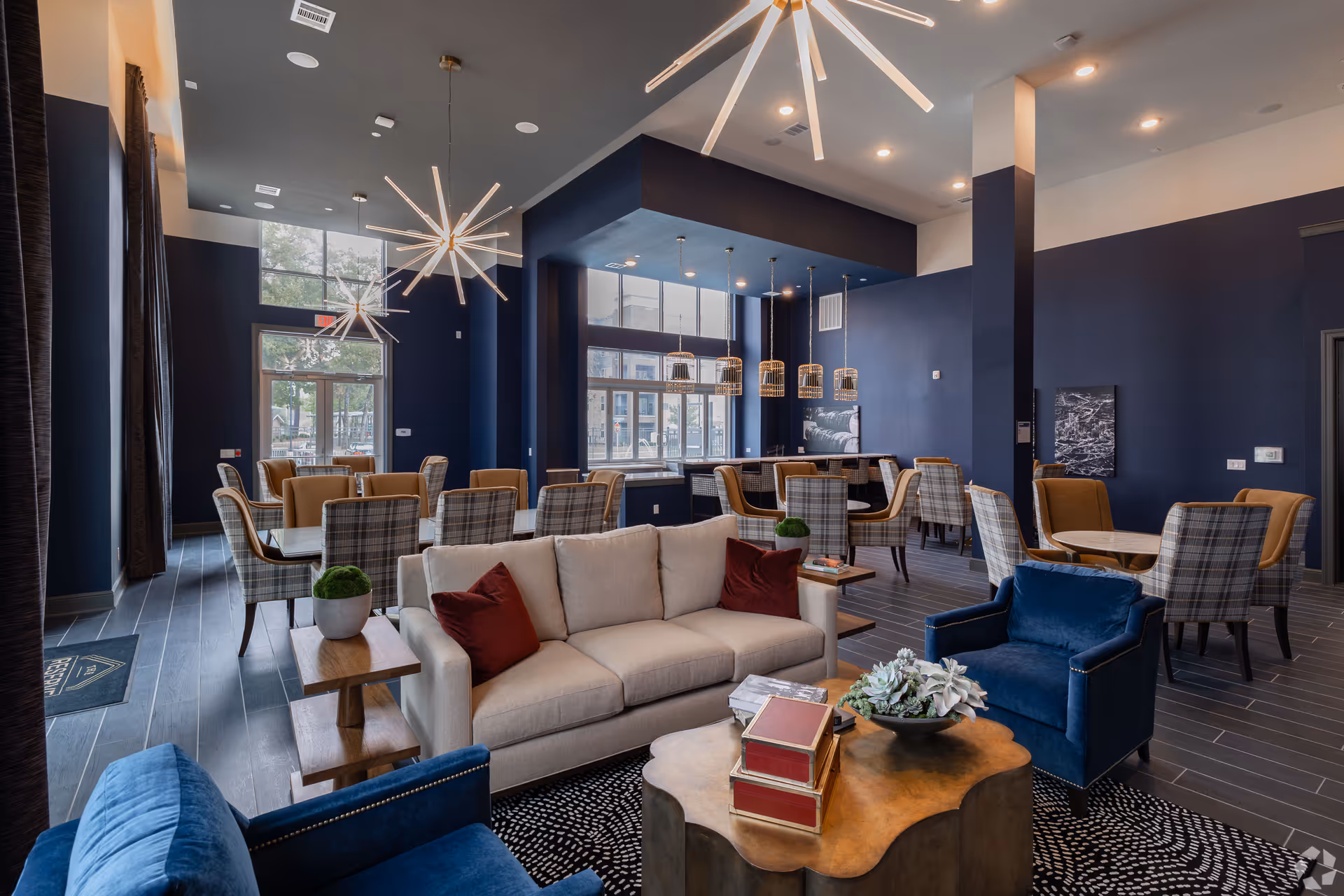 A modern and stylish common area with a beige sofa, blue armchairs, and a wooden coffee table with decorative books and a plant. The room features multiple dining tables with plaid and tan chairs, large windows letting in natural light, dark blue walls, and contemporary pendant lighting fixtures hanging from the ceiling.