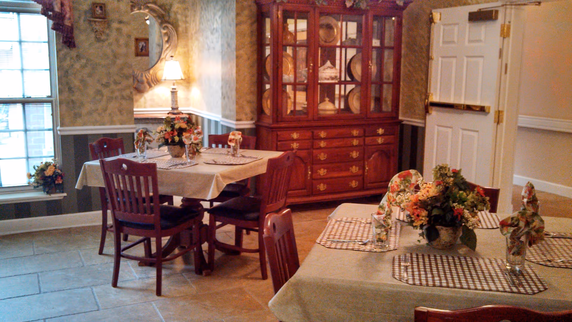 A cozy dining room with two tables covered in beige tablecloths, each set with checkered placemats, floral napkins in glasses, and a centerpiece of colorful flowers. The room features wooden chairs, a large wooden china cabinet filled with decorative plates, a wall lamp with a mirror above it, and a window letting in natural light.
