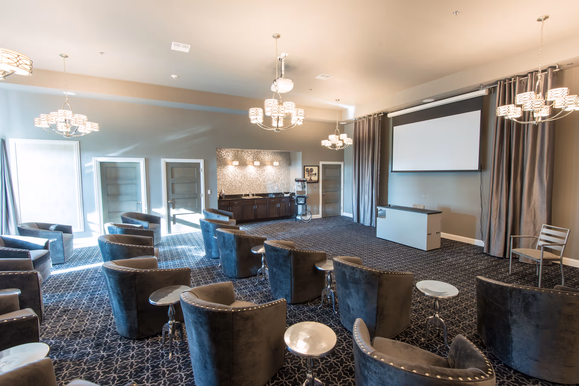 A spacious, well-lit meeting or presentation room with multiple gray upholstered chairs arranged in rows facing a large projection screen. The room features patterned carpet, modern chandeliers, a sideboard with decorative lighting, and several small round tables between chairs. There are three closed doors and large windows with curtains allowing natural light to enter.