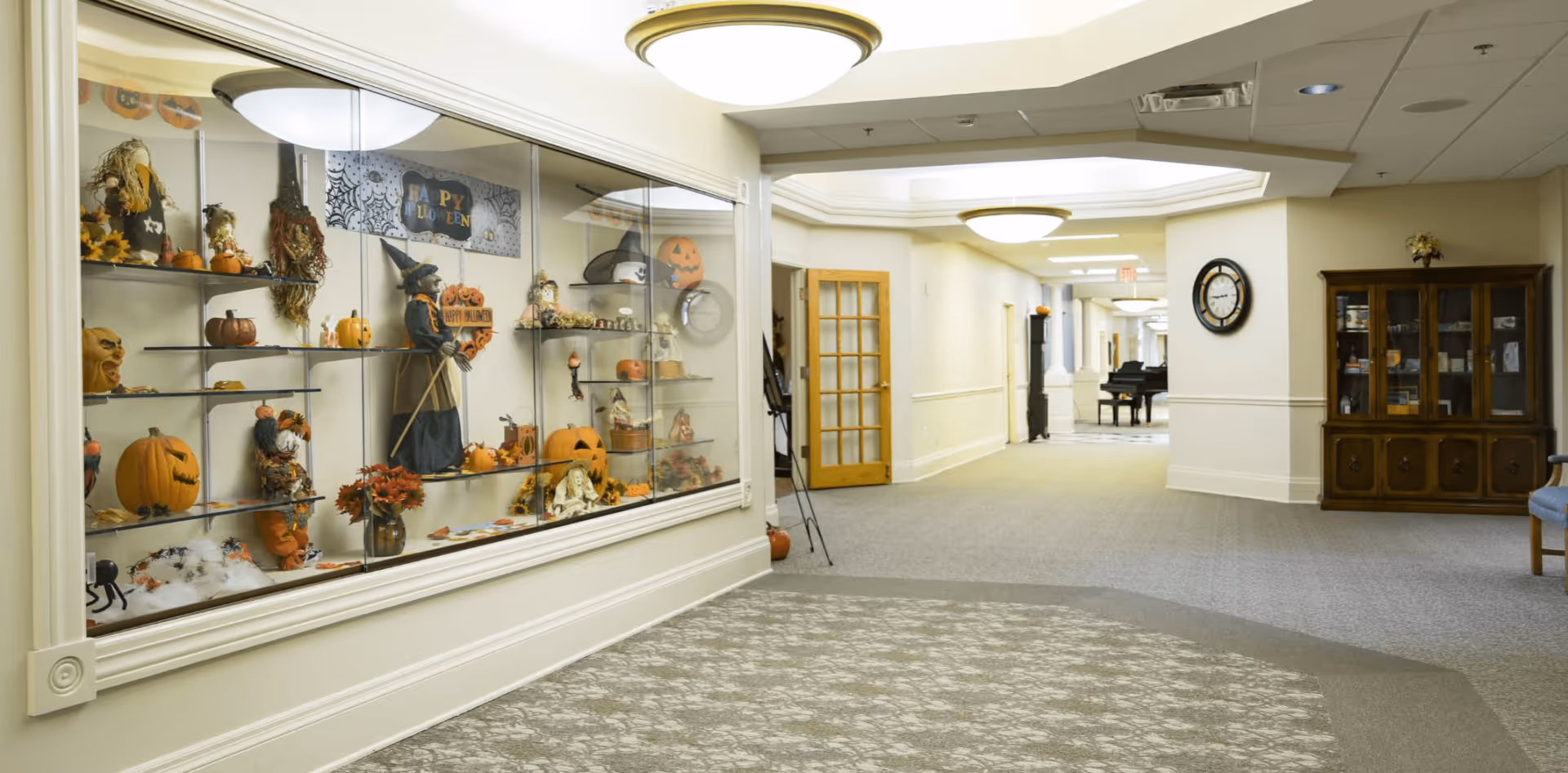 Interior hallway of a senior living facility decorated for Halloween with a glass display case filled with pumpkins, scarecrows, and Halloween-themed decorations. The hallway has beige walls, carpeted floors, ceiling lights, a wooden cabinet with glass doors, a wall clock, and a piano in the distance.