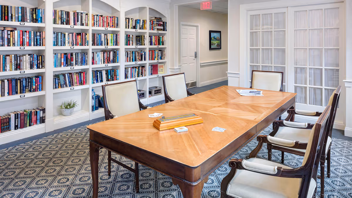 A well-lit room with a large wooden table surrounded by six cushioned chairs. Behind the table is a wall of built-in white bookshelves filled with books. The floor is covered with a patterned carpet, and there are French doors with sheer curtains on one side of the room.