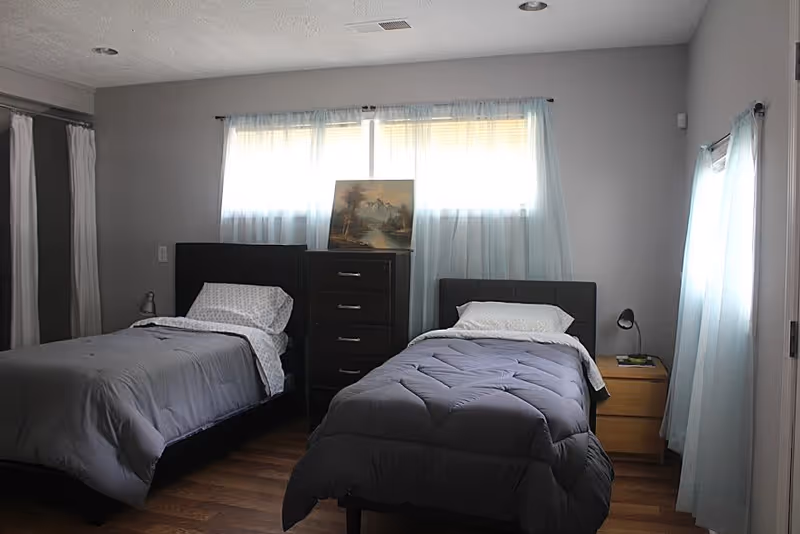 A bedroom with two single beds, each with gray bedding and white pillows. Between the beds is a dark wooden chest of drawers with a painting resting on top. There is a small wooden nightstand with a lamp and a book next to the bed on the right. The room has light gray walls, wooden flooring, and windows with sheer white curtains allowing natural light to enter.