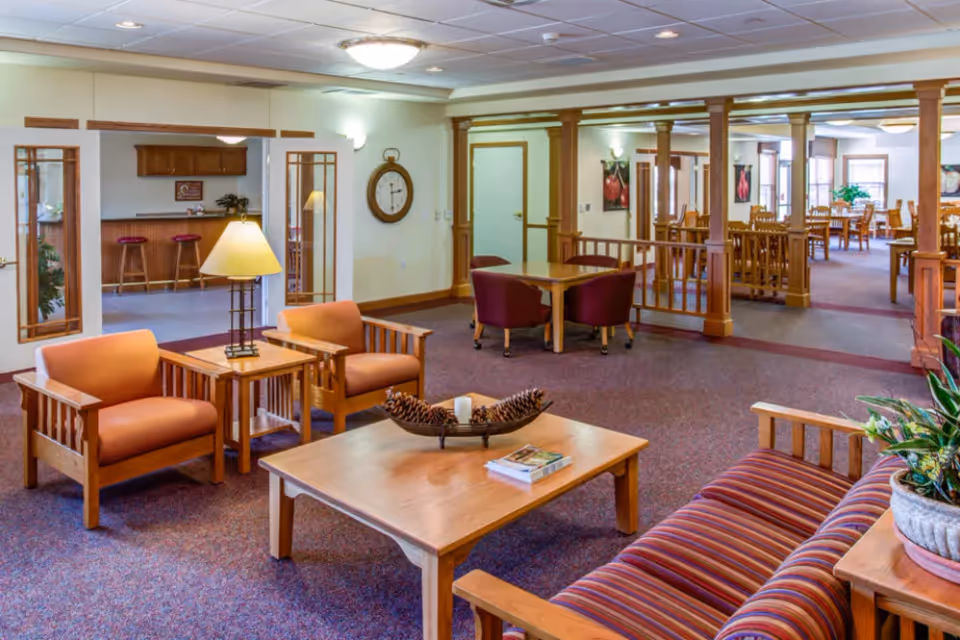 A spacious senior living facility common area with wooden furniture including a striped sofa, two armchairs, a coffee table with decorative pinecones and a candle, and a side table with a lamp. In the background, there are dining tables and chairs, wooden columns, and a wall clock. The room has carpeted floors and soft lighting.