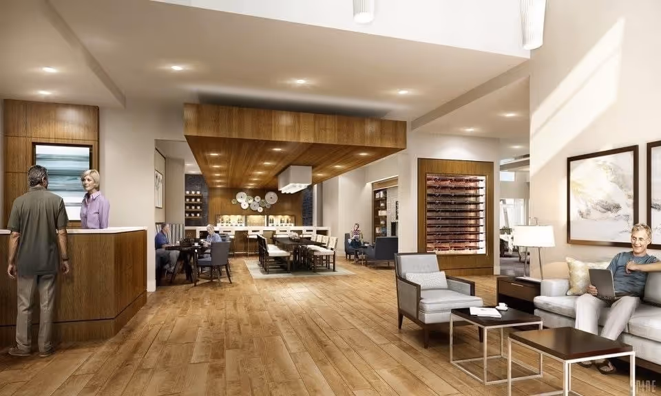 A spacious and modern senior living facility common area with wooden flooring and ceiling accents. On the left, a man is talking to a woman behind a reception desk. In the middle, several seniors are seated at tables, engaging in conversation. On the right, a man is sitting on a sofa with a laptop, next to a coffee table and armchair. The room is well-lit with recessed lighting and natural light from windows, and decorated with framed artwork and a wine rack.