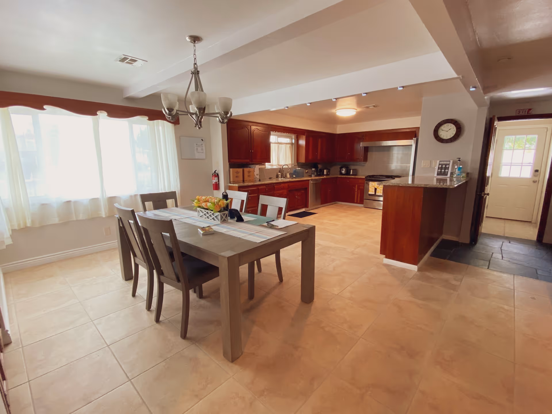 A spacious kitchen and dining area with a wooden dining table and six chairs. The kitchen features wooden cabinets, a stainless steel stove, and a dishwasher. There is a large window with white curtains letting in natural light, a chandelier above the dining table, and a clock on the wall near the entrance door.
