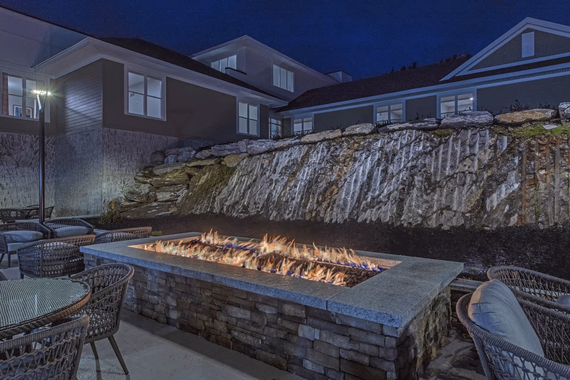 Outdoor patio area at night with a rectangular stone fire pit burning flames, surrounded by wicker chairs with cushions and a glass-top table. Behind the fire pit is a rock retaining wall and a building with multiple windows illuminated from inside.