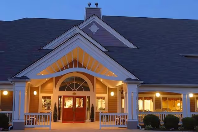 Front entrance of a warmly lit senior living facility with a covered porch, double red doors, columns, and surrounding windows at dusk.