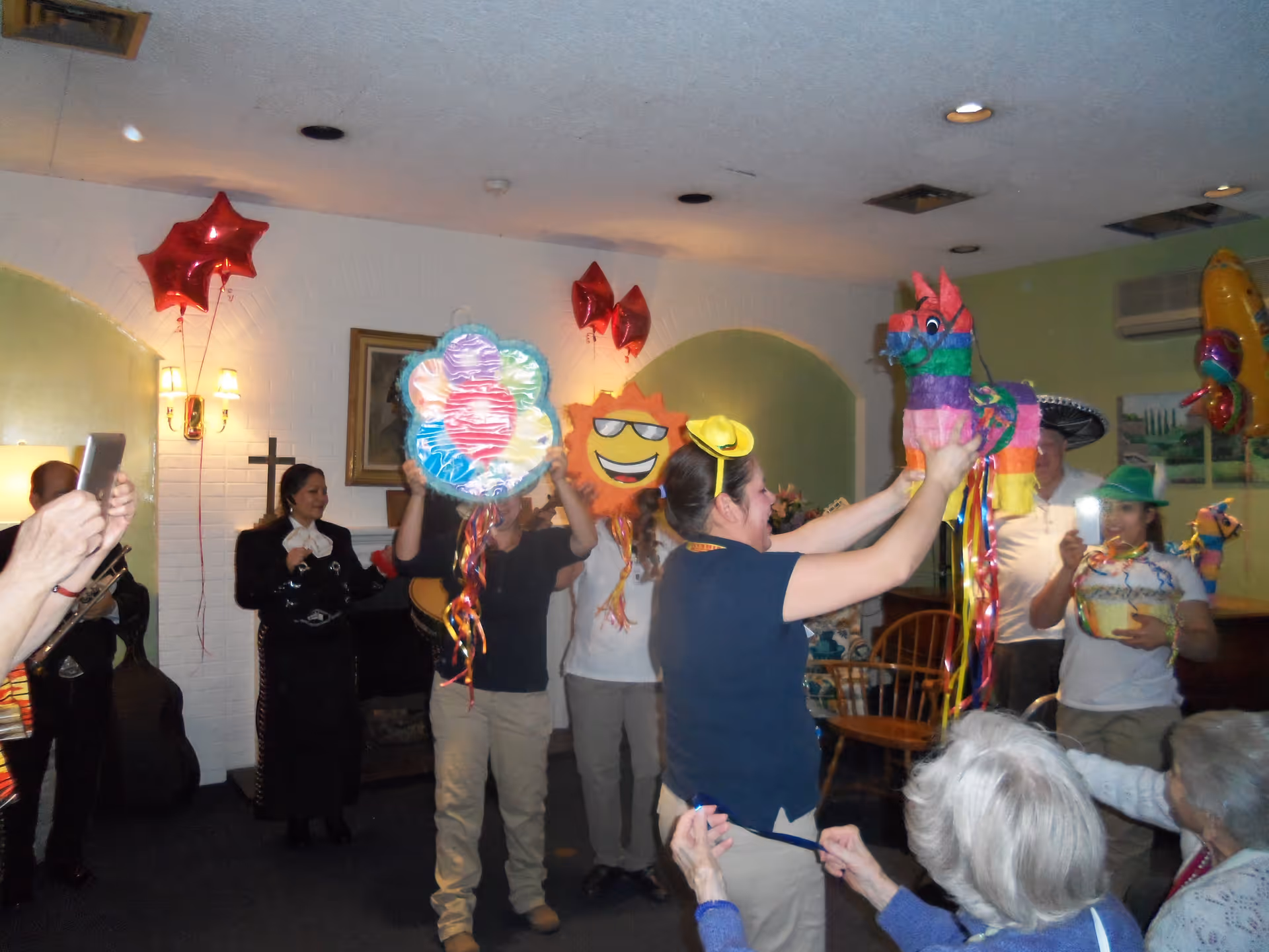 A group of people indoors celebrating with colorful piñatas shaped like a flower, a sun with sunglasses, and a donkey. Some people are wearing festive hats, and there are red star-shaped balloons attached to the wall. Two elderly individuals are seated in the foreground watching the celebration.
