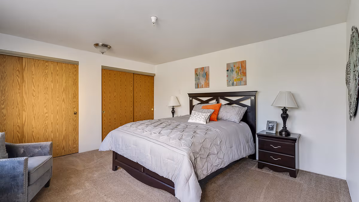 A neatly made bed with gray bedding and decorative pillows in a bedroom. There are two wooden sliding closet doors on the left wall, a dark wooden nightstand with a lamp and a framed photo on the right side of the bed, and two abstract paintings hanging above the bed. A gray armchair is partially visible on the left side of the image.
