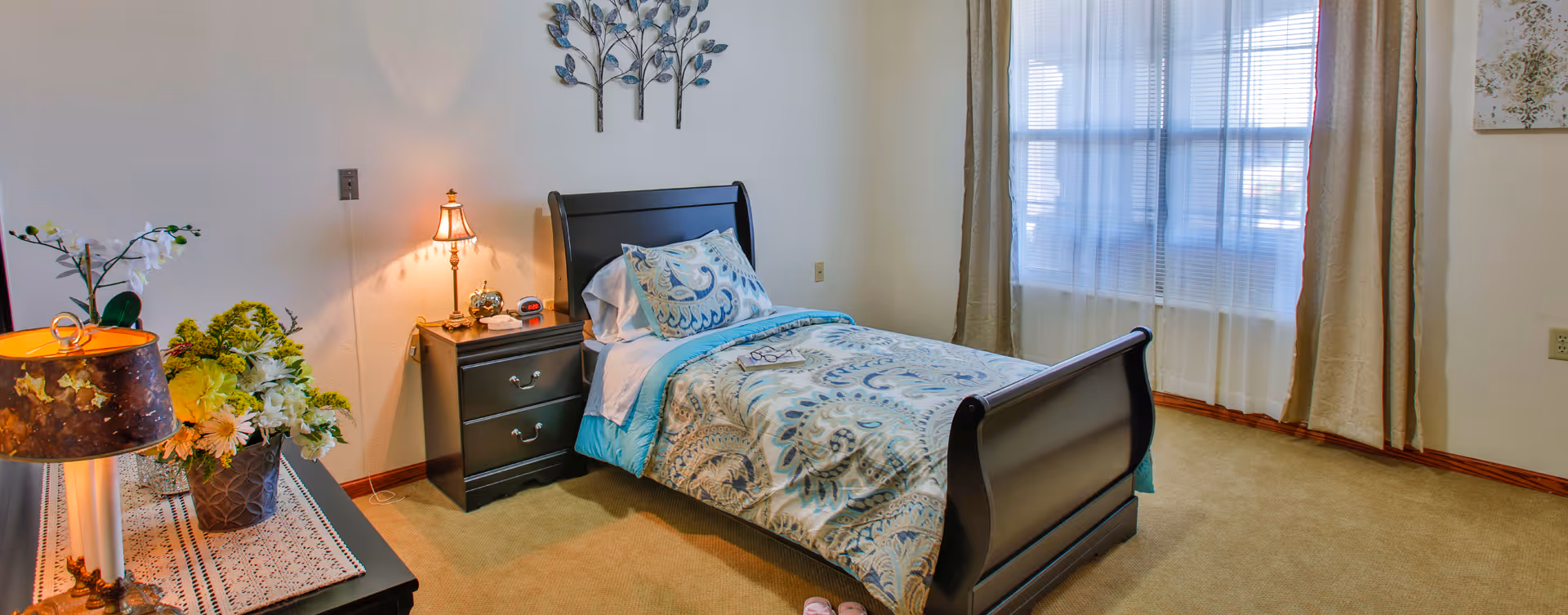 A cozy bedroom with a single bed featuring a blue and beige patterned comforter and pillow. Next to the bed is a dark wooden nightstand with a small lamp, an alarm clock, and a decorative item. A large window with sheer white curtains and beige drapes allows natural light to fill the room. A dresser with a lamp and flower arrangements is visible on the left side. The walls are light-colored, and there is a decorative metal wall art piece above the bed.