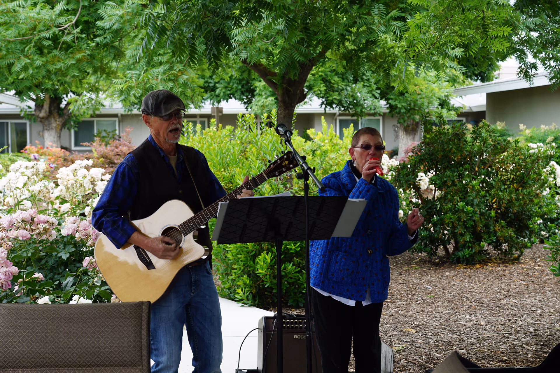 An elderly man playing an acoustic guitar and singing into a microphone while an elderly woman stands beside him drinking from a cup in a garden area with trees and flowering bushes.