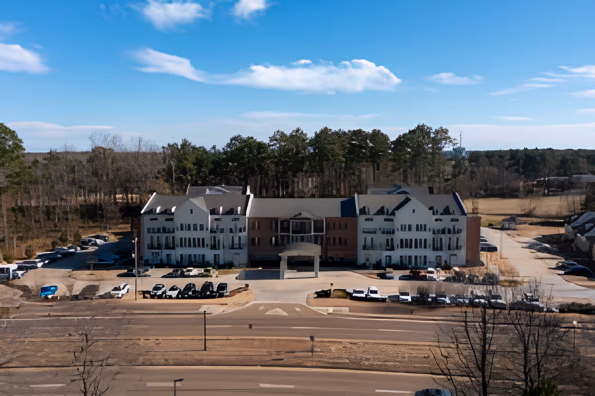 Front exterior of a multi-story senior living facility with a parking lot, surrounding trees, and a blue sky.