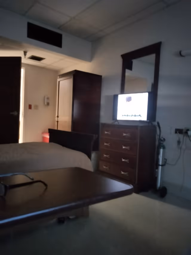 A dimly lit room with a bed, a wooden dresser with a mirror, a television on top of the dresser, an oxygen tank beside the dresser, a wooden wardrobe, and a table with a pair of glasses on it. The door to the room is open, letting in some light.