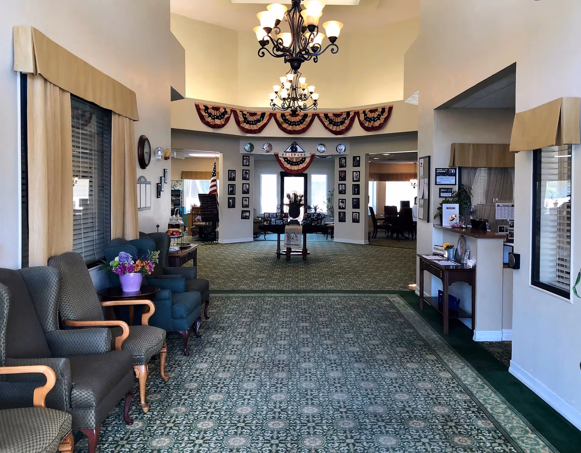 Interior view of a senior living facility lobby area with green patterned carpet, several upholstered chairs along the left wall, a small table with a purple flower pot, and a reception desk on the right. The far end features a decorated wall with patriotic bunting, framed photos, and a sign that reads 'Veterans'. Two chandeliers hang from the ceiling.